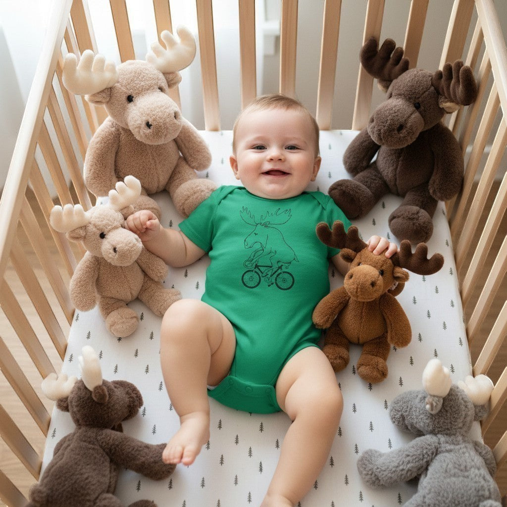 Rack & Cycle green cotton baby bodysuit with moose on bicycle graphic, surrounded by plush moose toys in a crib.